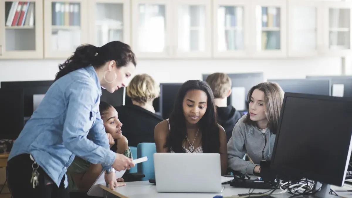 A female school teacher in a computer lab speaking to three teenage girls sitting in front of a laptop. 