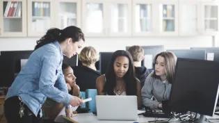 A female school teacher in a computer lab speaking to three teenage girls sitting in front of a laptop. 