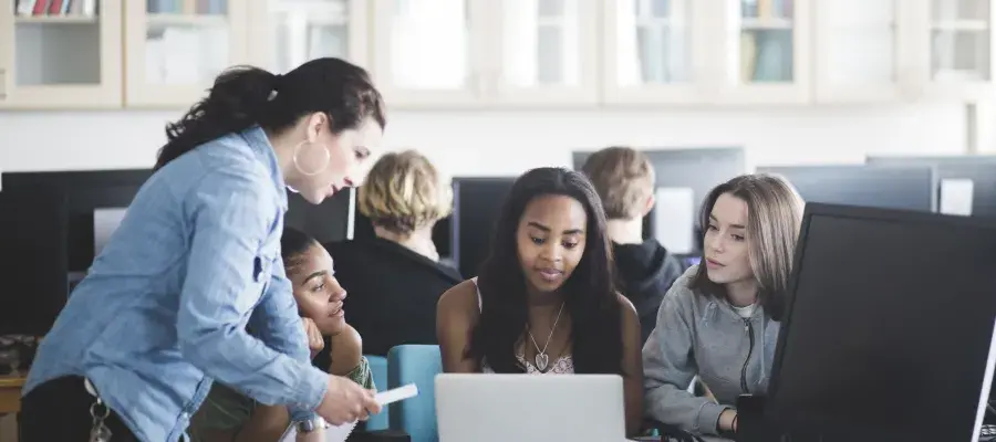 A female school teacher in a computer lab speaking to three teenage girls sitting in front of a laptop. 