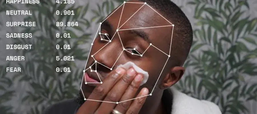 A young black man looks into the camera. He is wearing a dressing gown and has a towel around his neck. He applies a cotton pad to his face and wears a ring on his right hand. a list of words and numbers describe the man’s expression. Happiness 4.185, neutral 0.901, surprise 89.864, sadness 0.01, disgust 0.01, anger 5.021 and fear 0.01.