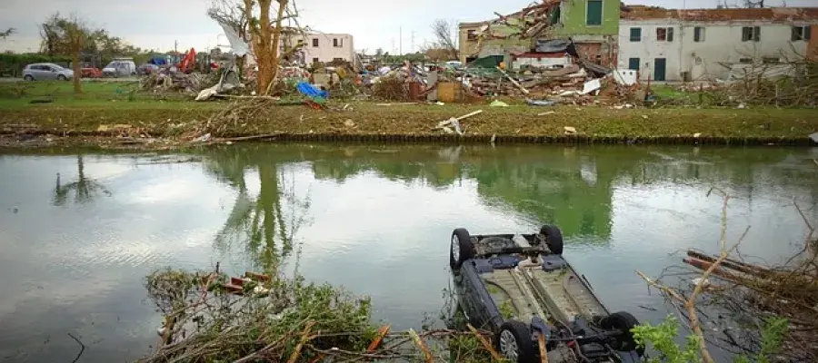 A picture of the aftermath of a torndado, in the background there is a building on the left side that has been ripped in half. In the middle of the background is a tree that has been damaged. The middle ground has flooding. The foreground has a car that has been turned upside down. 