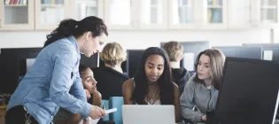 A female school teacher in a computer lab speaking to three teenage girls sitting in front of a laptop. 