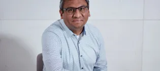 A photograph of Professor Subramanian Ramamoorthy sitting at a desk with his right elbow on the desk.  a desk with 