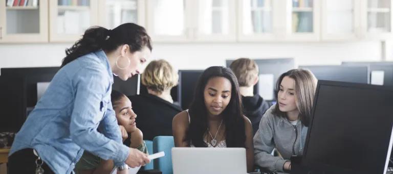 A female school teacher in a computer lab speaking to three teenage girls sitting in front of a laptop. 