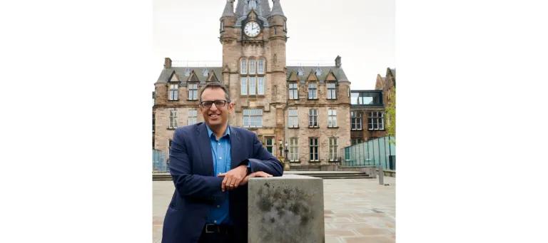 A photograph of Kev Dhaliwal wearing a suit, leaning against a pillar standing in front of the Edinburgh Futures Institute. 