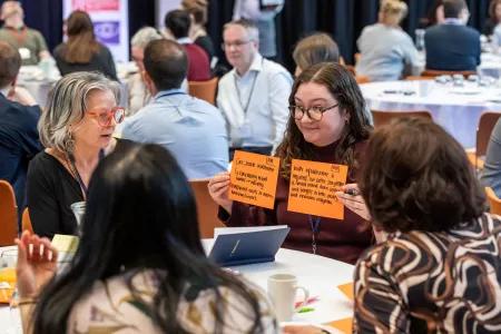 A woman wearing glasses sitting at a table holds up two flash cards to three other people at table outlining details of a discussion that they have just had about AI infrastructure. 
