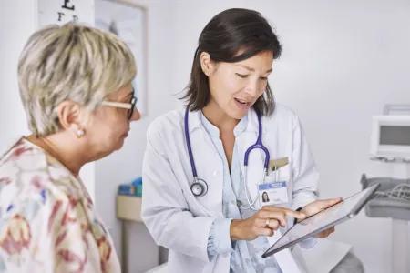 Female doctor explaining electronic test results to female patient at clinic. They are sitting in examination room and the doctor has an ipad. The doctor is wearing a white coat and has a stethoscope around her neck. The patient is wearing a floral shirt and glasses.