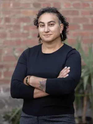 A portrait photograph of Dr Alex Hanna, standing facing the camera with her arms folded across her chest. 