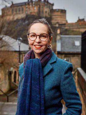 A portrait photograph of Professor Shannon Vallor smiling with a scarf on and big coat standing at the top of some steps with Edinburgh Castle in the background. 