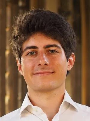 A photo of Edoardo Point who has short brown hair and is wearing a white shirt, he is standing in from of a gold background. 