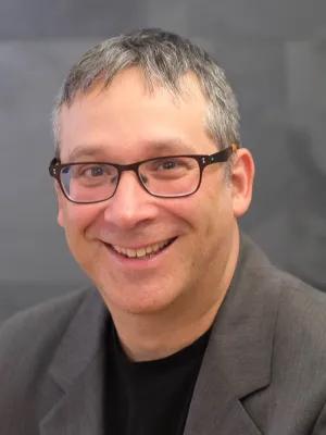 A picture of a white man with glasses and grey hair, you only see the top half of his body and he is front of a grey backgtound. He is wearing a grey blazer and black shirt. 